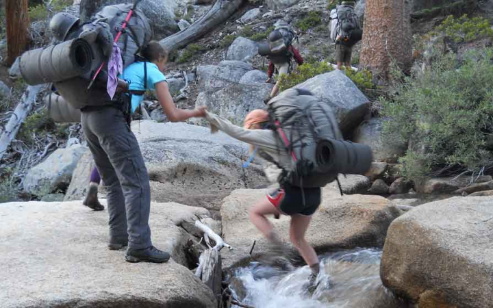 One backpacker helps another cross a rocky stream.